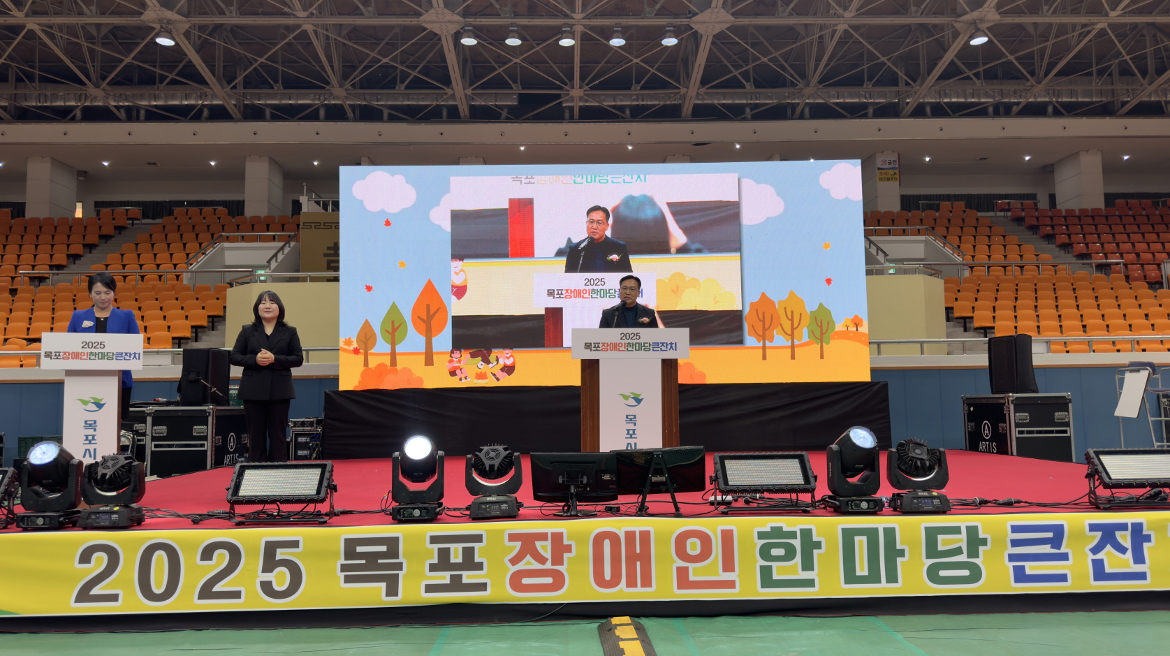 실내 무대 중앙 연단에서 조석훈 목포시 부시장이 2025 목포장애인한마당큰잔치 개회 인사말을 하고 있고, 뒤 전광판에는 가을 숲 배경 그래픽과 연단 모습이 함께 비치며 왼쪽에는 사회자와 수어 통역사가 서 있는 장면.