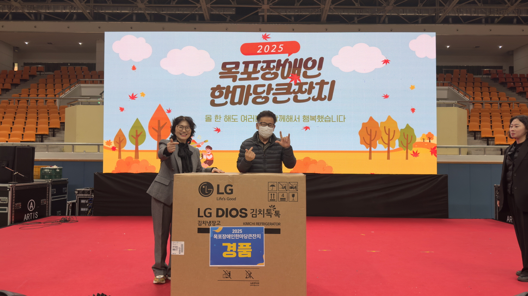 2025 목포장애인한마당큰잔치 무대 중앙에서 ‘경품’ 스티커가 붙은 대형 LG 김치냉장고 상자 앞에 추첨에 당첨된 참가자와 진행자가 서서 엄지손가락을 치켜세우며 환하게 웃고 있고, 뒤 전광판에는 행사 로고와 ‘올 한 해도 여러분과 함께해서 행복했습니다’라는 문구가 보이는 모습.