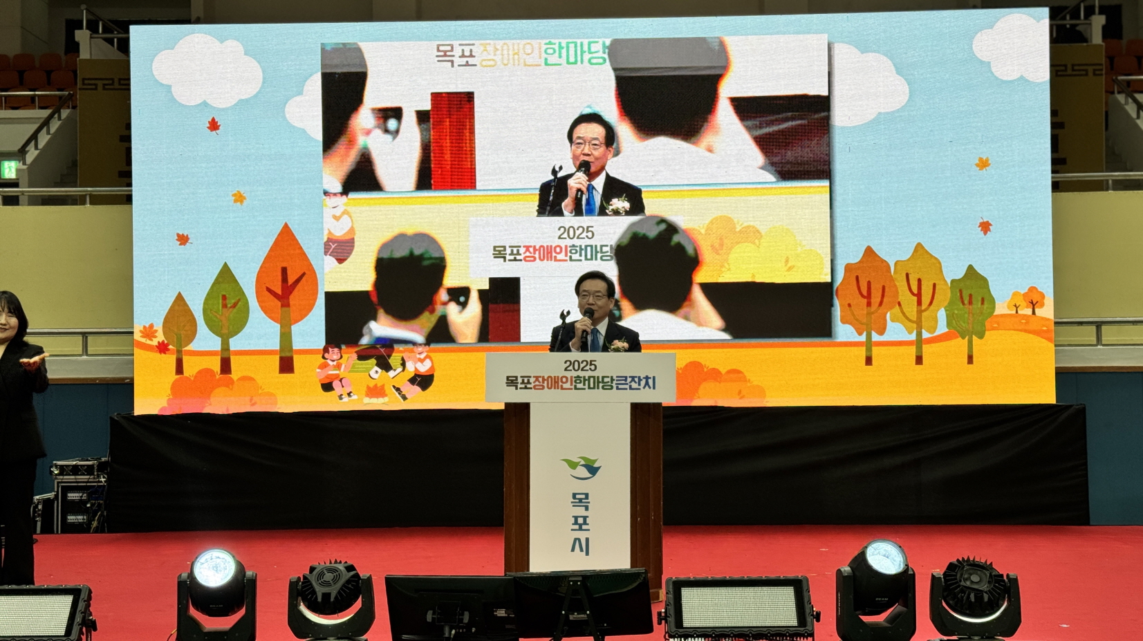 가을 숲 그림이 배경으로 나오는 대형 전광판 앞 연단에서 조성오 목포시의회 의장이 2025 목포장애인한마당큰잔치 축사를 하고 있고, 전광판에는 의장의 발언 모습이 크게 비치고 연단 아래에는 ‘2025 목포장애인한마당큰잔치’ 문구가 적힌 모습.