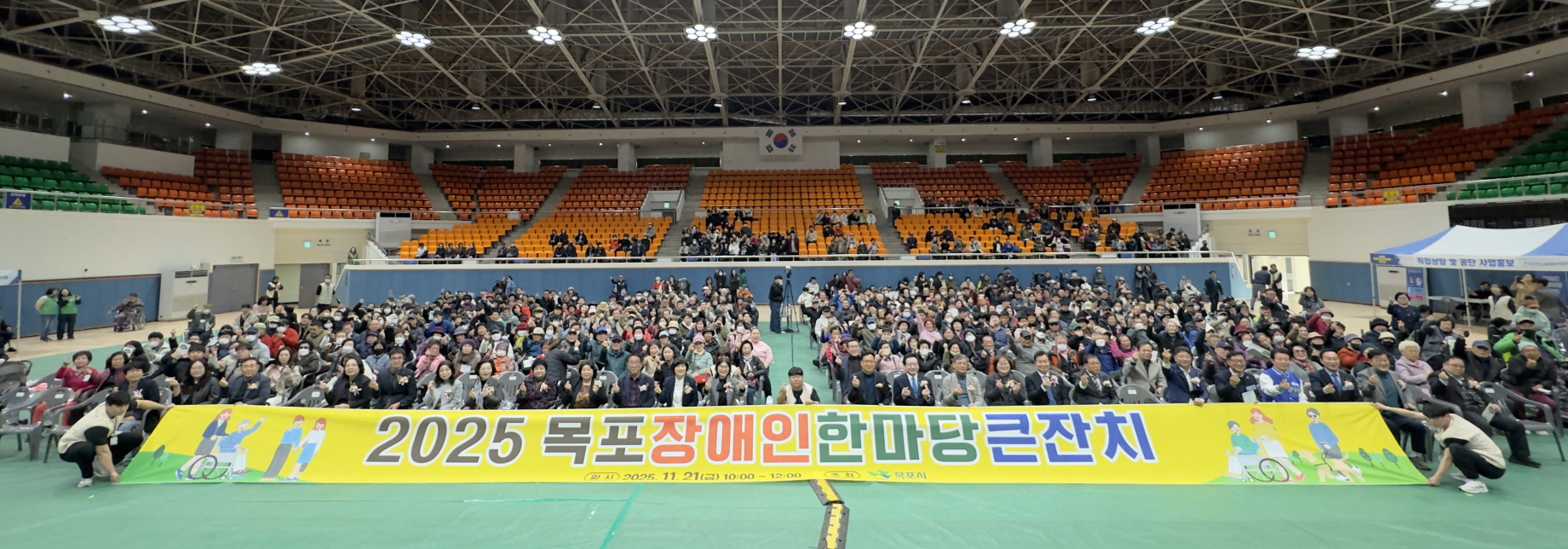 실내 체육관 관중석을 배경으로, 바닥 중앙에 ‘2025 목포장애인한마당큰잔치’라고 적힌 노란 현수막을 펼쳐 들고 장애인과 가족, 시민 수백 명이 함께 앉아 카메라를 향해 손하트를 하며 기념촬영하고 있는 모습.