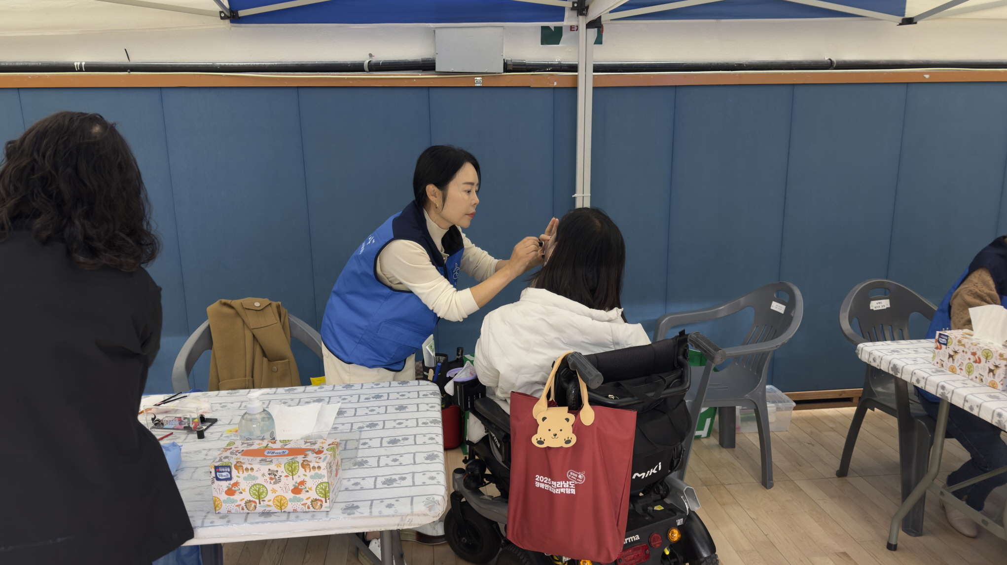 실내 부스 천막 아래에서 ‘목포를 만드는 사람들’ 봉사자가 휠체어를 이용하는 참가자의 얼굴에 세심하게 페이스페인팅을 해 주는 모습.