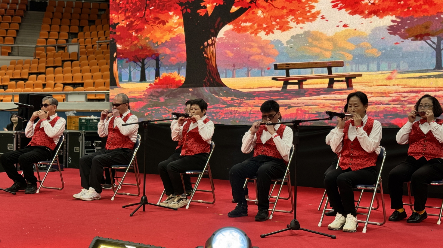 가을 단풍이 물든 공원을 배경으로 한 전광판 앞에서 한국시각장애인연합회 목포지회 이용고객들이 빨간 조끼와 흰 셔츠를 맞춰 입고 의자에 앉아 하모니카를 합주하는 2025 목포장애인한마당큰잔치 무대 공연 장면.