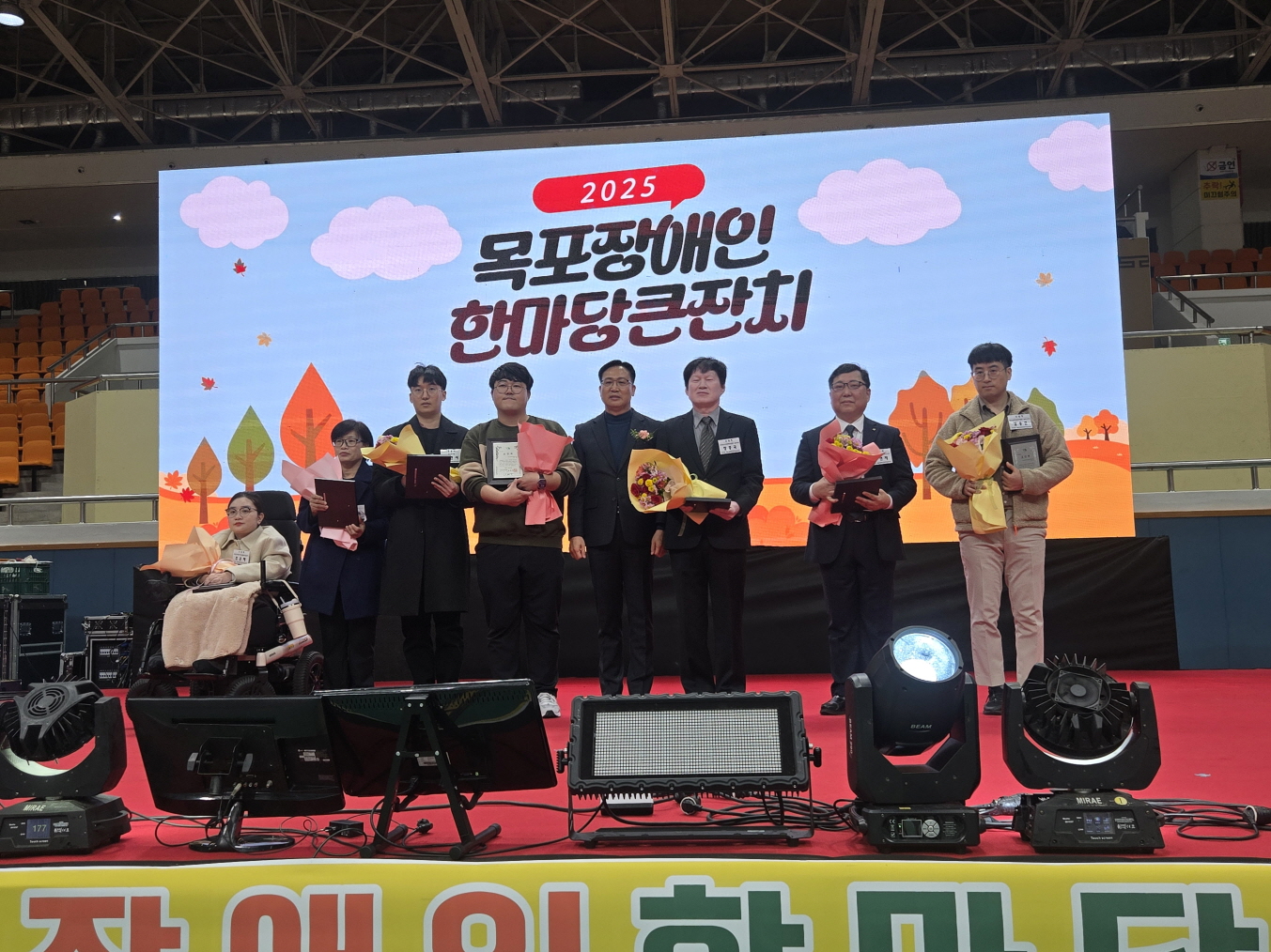 2025 목포장애인한마당큰잔치 무대 위에서 장애인과 자원봉사자, 기관 관계자들이 나란히 서서 공로패와 꽃다발을 들고 기념촬영하고 있으며, 뒤 전광판에는 행사 로고와 ‘목포장애인한마당큰잔치’라는 글자가 보이는 시상 장면.