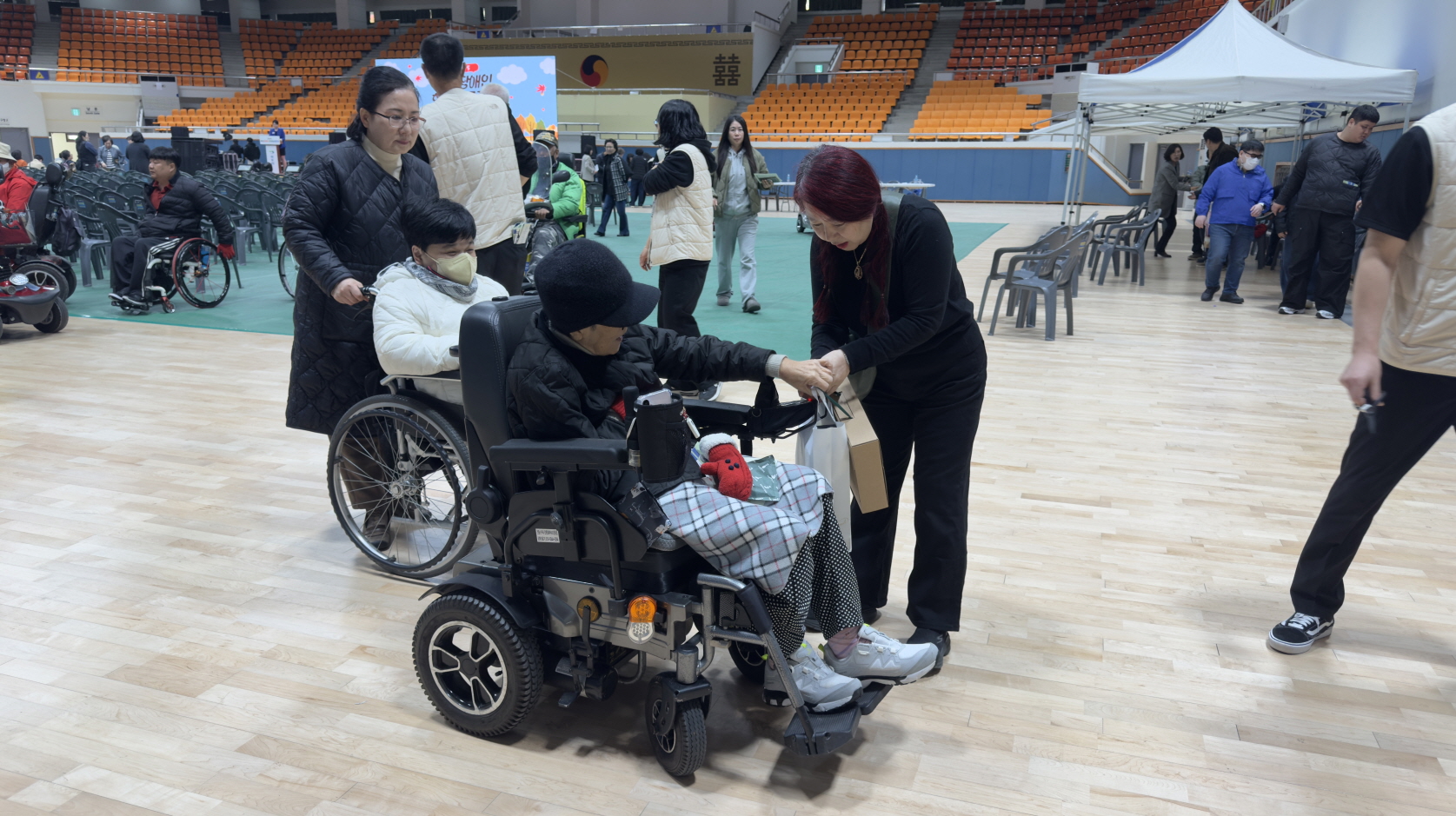 행사장을 나가며 전동휠체어를 이용하는 참가자가 이동하고 있고, 검은 옷을 입은 스태프가 두 손으로 기념품 봉투를 건네며 인사를 나누고 있으며, 주변에는 다른 휠체어 이용고객과 보호자가 함께 퇴장 준비를 하는 모습.