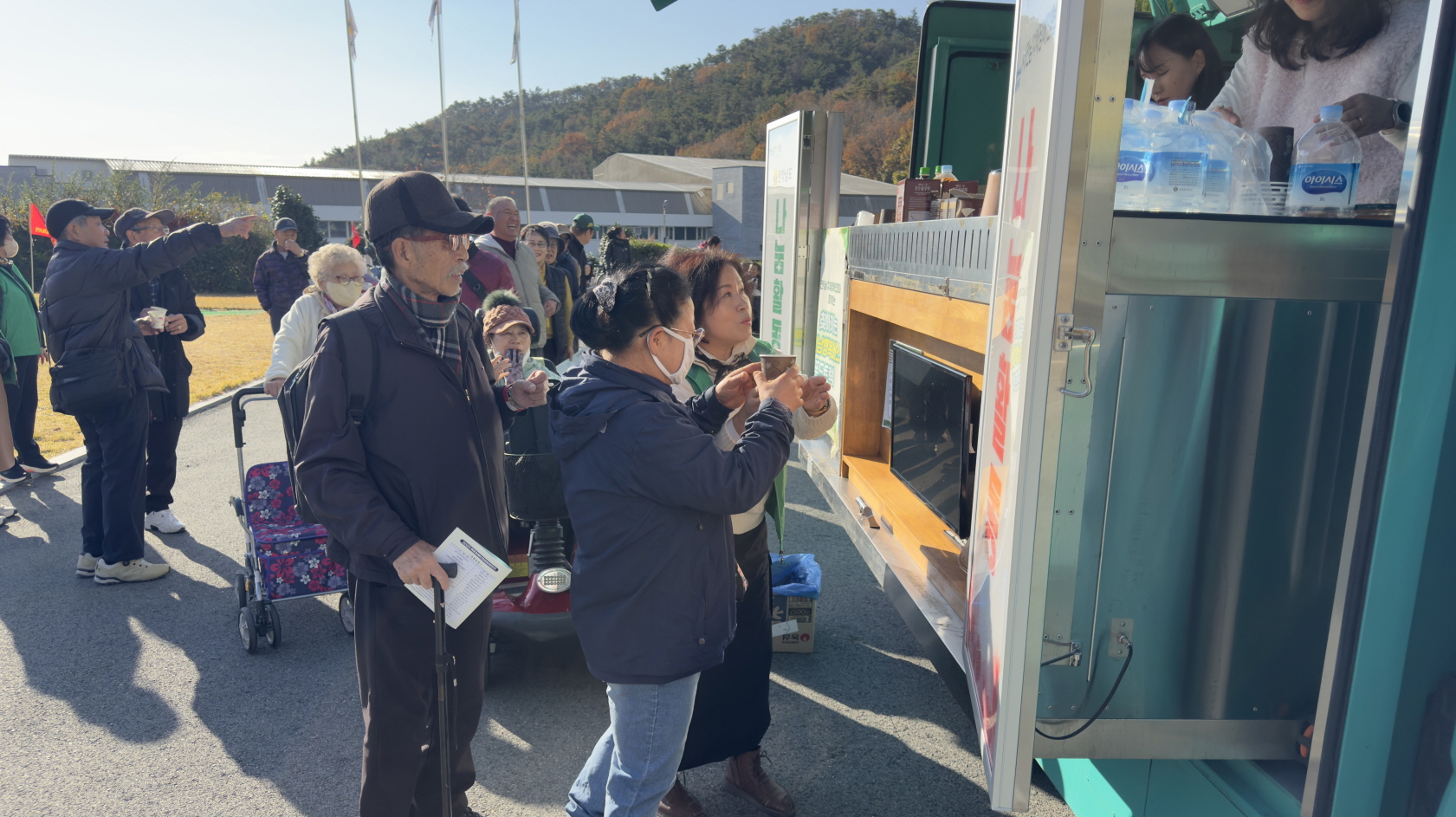 야외에 주차된 푸드트럭 앞에서 어르신과 장애인 당사자들이 줄을 서서 따뜻한 음료와 간식을 무료로 받으며 대화를 나누고 있고, 뒤쪽으로는 맑은 하늘과 단풍든 나무가 보이는 모습.