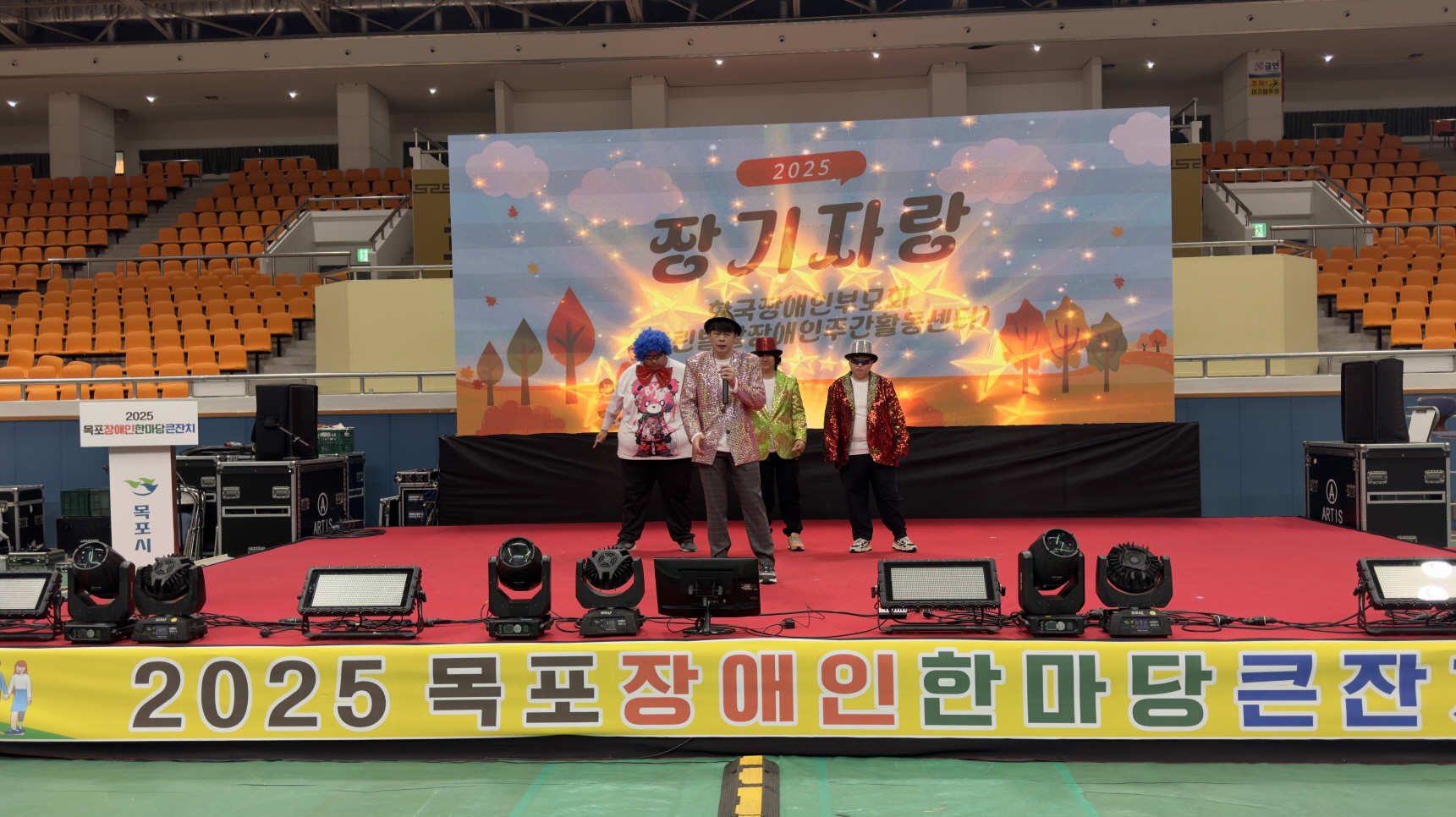 실내 무대 위에서 한국장애인부모회 열린발달장애인주간활동센터 이용고객들이 반짝이는 의상과 모자를 착용하고 기관 공연으로 노래와 댄스를 선보이고 있으며, 뒤 전광판에는 ‘장기자랑’ 문구와 화려한 그래픽이 나타난 모습.