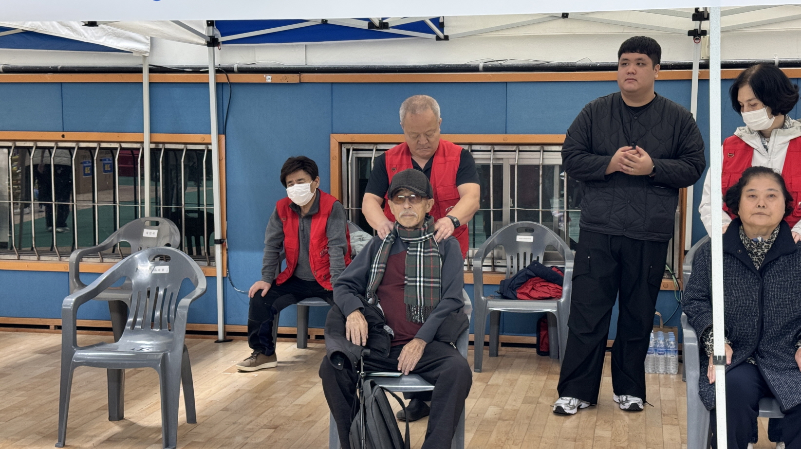 실내 체육관 한쪽 건강관리 부스에서 전라남도시각장애인협회 목포지부 안마사가 의자에 앉은 어르신의 어깨와 목을 시원하게 주무르며 시각장애인 안마 체험을 제공하는 장면.
