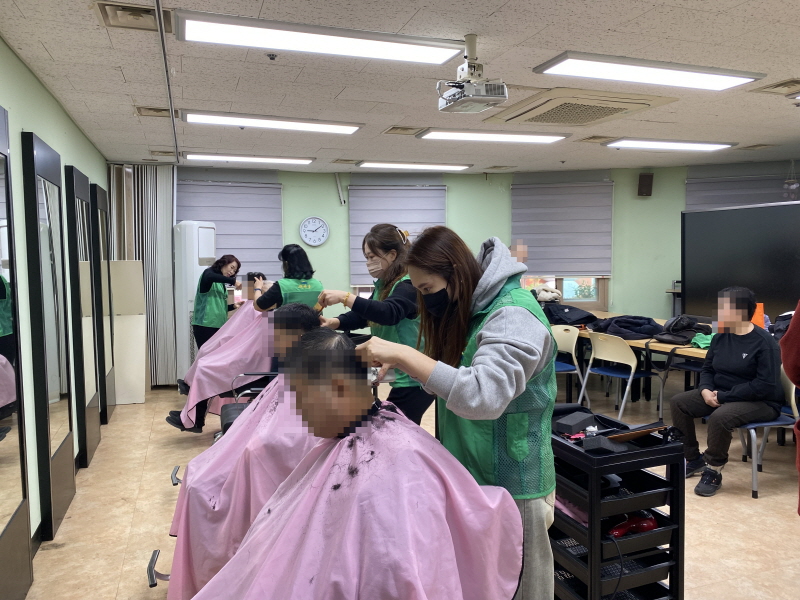 앞쪽에서 초록색 조끼와 회색 후드티를 입은 하당동 새마을부녀회 미용봉사자가 남성 이용고객의 머리를 꼼꼼히 자르고, 뒤편 거울 앞에서도 다른 부녀회원들이 이미용 서비스를 제공하며 오른쪽 의자에는 차례를 기다리는 이용고객들이 앉아 있는 모습
