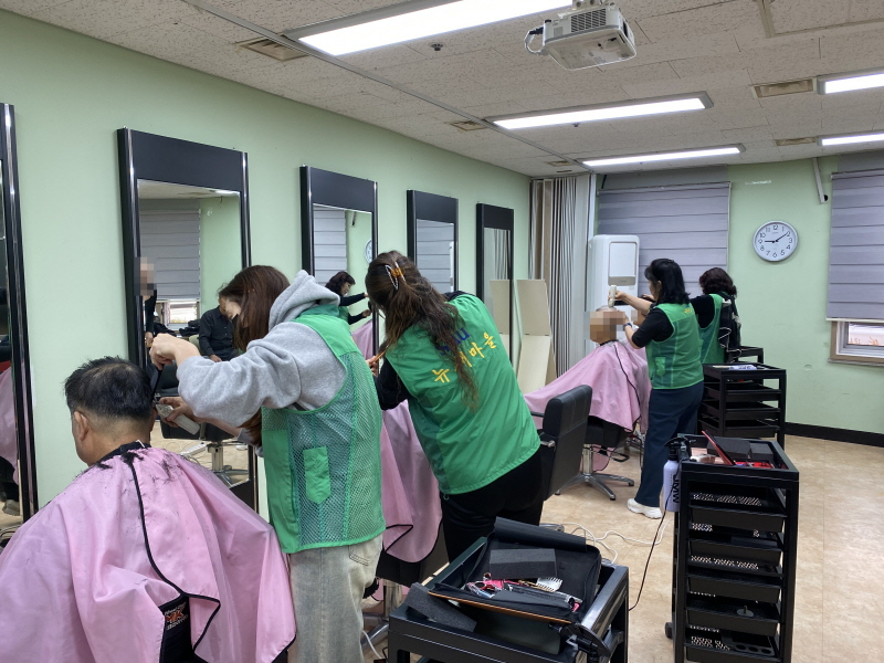 초록색 조끼를 입은 하당동 새마을부녀회 봉사자들이 거울 앞에 서서 분홍색 커트보를 두른 남성 이용고객들의 머리를 동시에 다듬고 있으며, 미용 도구 카트가 줄지어 놓인 복지관 이미용실 풍경