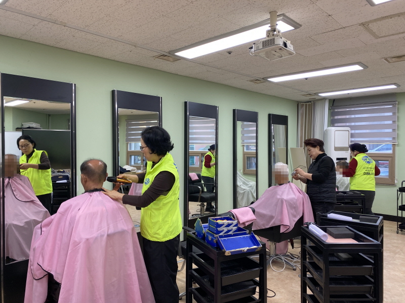 여러 개의 거울 앞에서 노란 조끼를 입은 병아리봉사단 봉사자와 검은 옷의 미용사가 분홍색 커트보를 두른 남성 이용고객들의 머리를 손질하고 있으며, 작업대와 미용 도구가 정돈돼 있는 복지관 이미용실 전경