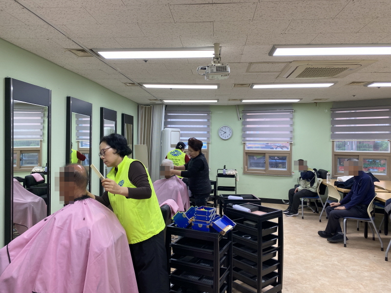 복지관 이미용실에서 노란 조끼를 입은 병아리봉사단 미용봉사자가 분홍색 커트보를 두른 남성 이용고객의 머리를 정성스럽게 자르고 있고, 오른쪽 의자에는 차례를 기다리는 이용고객들이 앉아 있는 모습