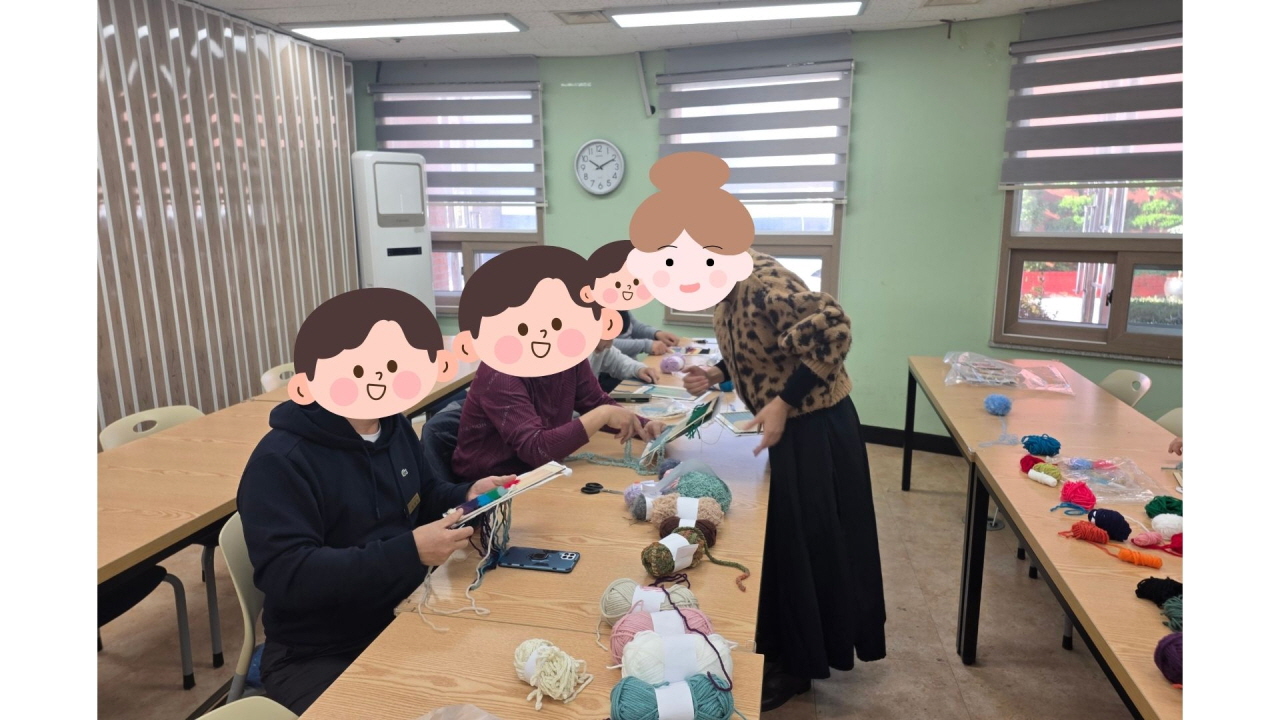 긴 책상에 나란히 앉은 성인 이용자들이 여러 색의 털실과 도구를 앞에 두고 펀치니들 기법으로 수를 놓고 있고, 강사가 각자의 손동작을 살펴보며 바늘 사용하는 방법을 자세히 지도해 주는 공예 수업 장면