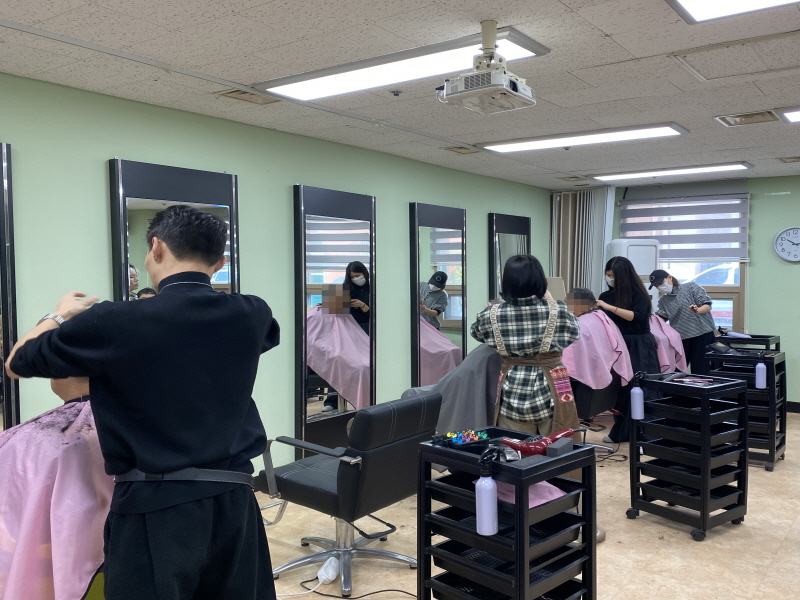 검은 작업복과 체크무늬 원피스를 입은 청담헤어 미용사들이 일렬로 놓인 의자에 앉은 남성 이용고객들의 머리를 동시에 자르고 있고, 각 자리 옆 카트에 빗과 가위, 스프레이 등이 놓인 활기찬 이미용 봉사 장면