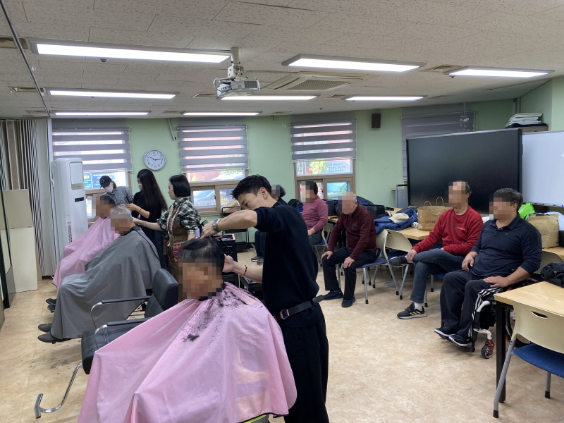 앞쪽에서는 분홍색과 회색 커트보를 두른 남성 이용고객들이 청담헤어 미용사에게 머리를 깎고 있고, 오른쪽 뒤편 줄지어 놓인 의자에는 차례를 기다리는 여러 남성 이용고객들이 앉아 있는 등 한 공간에서 이미용 서비스가 진행되는 모습