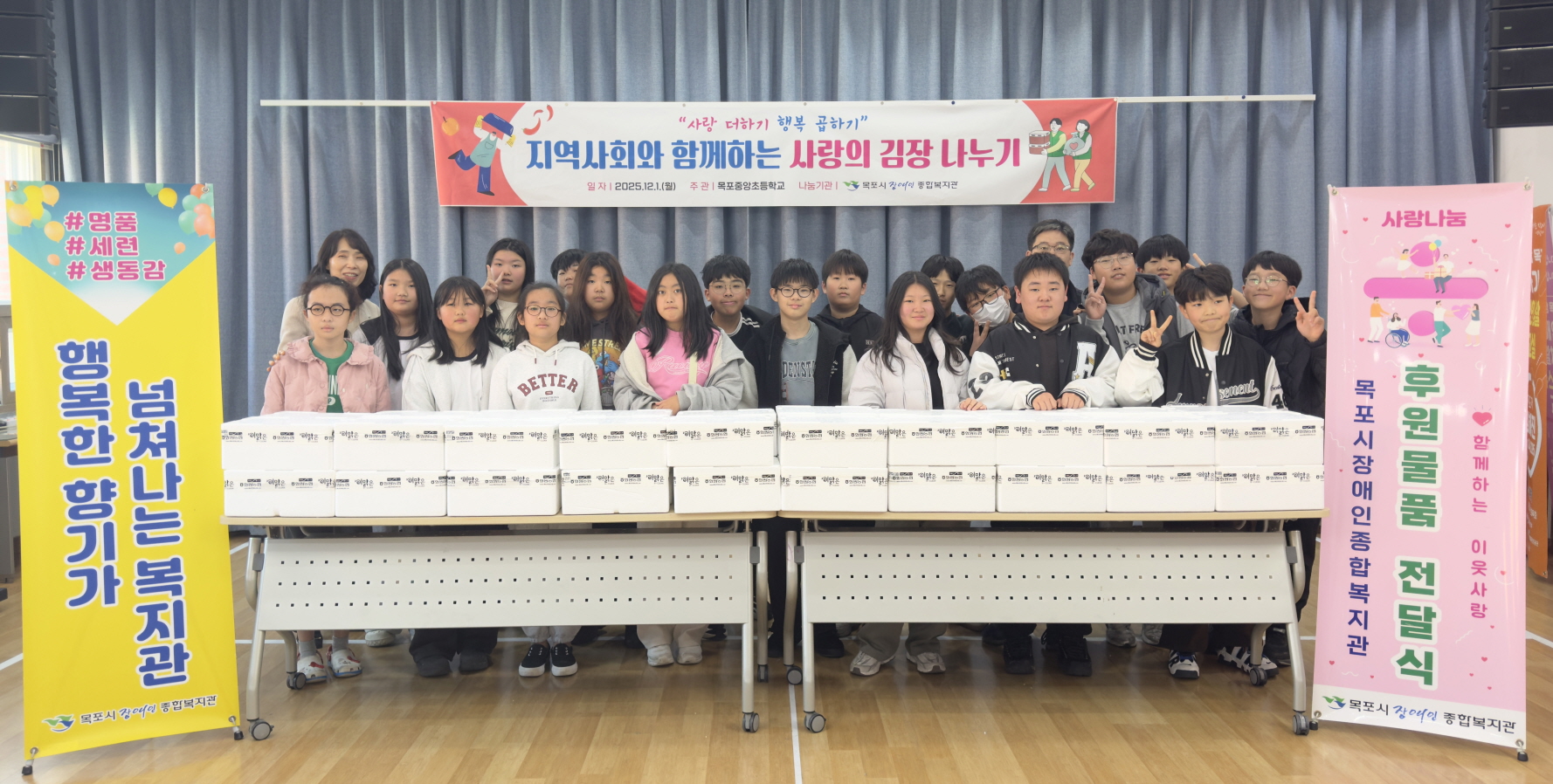 목포시장애인종합복지관 강당에서 열린 ‘지역사회와 함께하는 사랑의 김장 나누기’ 전달식에서 목포중앙초등학교 학생들이 김장 박스가 빼곡히 놓인 책상 뒤에 두 줄로 서 있다. 학생들은 일부는 주먹을 쥐고 일부는 V자를 그리며 환하게 웃고 있고, 뒤쪽에는 행사 제목이 적힌 현수막이, 양쪽에는 복지관을 소개하는 노란색·분홍색 배너가 세워져 있어 학교와 복지관이 함께 준비한 김장 나눔 후원물품 전달 현장임을 보여준다.