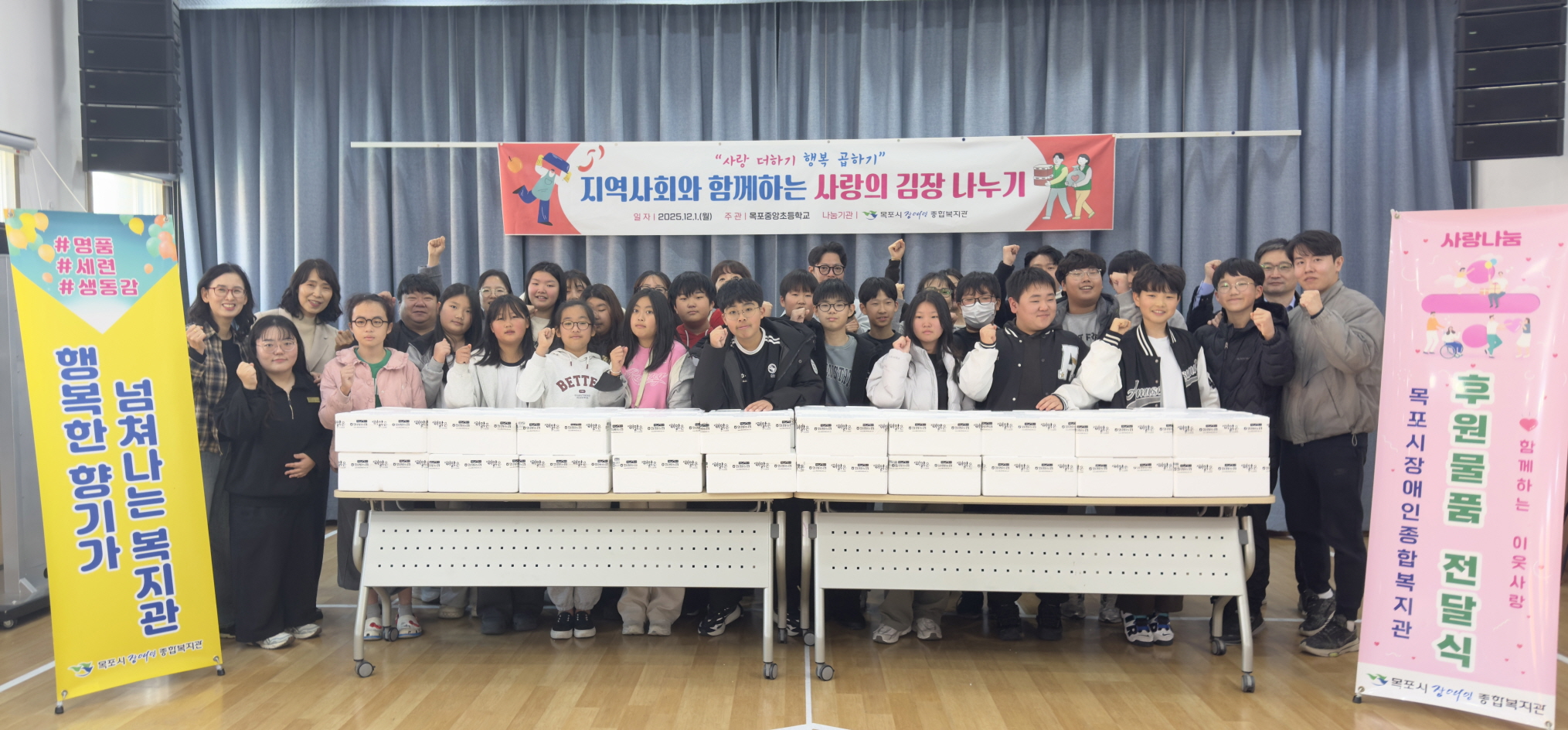 목포시장애인종합복지관 강당에서 ‘지역사회와 함께하는 사랑의 김장 나누기’ 전달식을 진행하며, 목포중앙초등학교 학생들과 교사, 복지관 관계자 등 30여 명이 김장 박스가 가득 쌓인 책상 뒤에 서서 카메라를 향해 주먹을 쥐고 응원의 포즈를 취하고 있다. 중앙 상단에는 행사 제목이 적힌 현수막이 걸려 있고, 좌우에는 ‘행복한 향기가 넘쳐나는 복지관’, ‘후원물품 전달식’이라고 적힌 세로형 배너가 세워져 있어 지역사회와 함께하는 나눔 활동임을 알 수 있다.