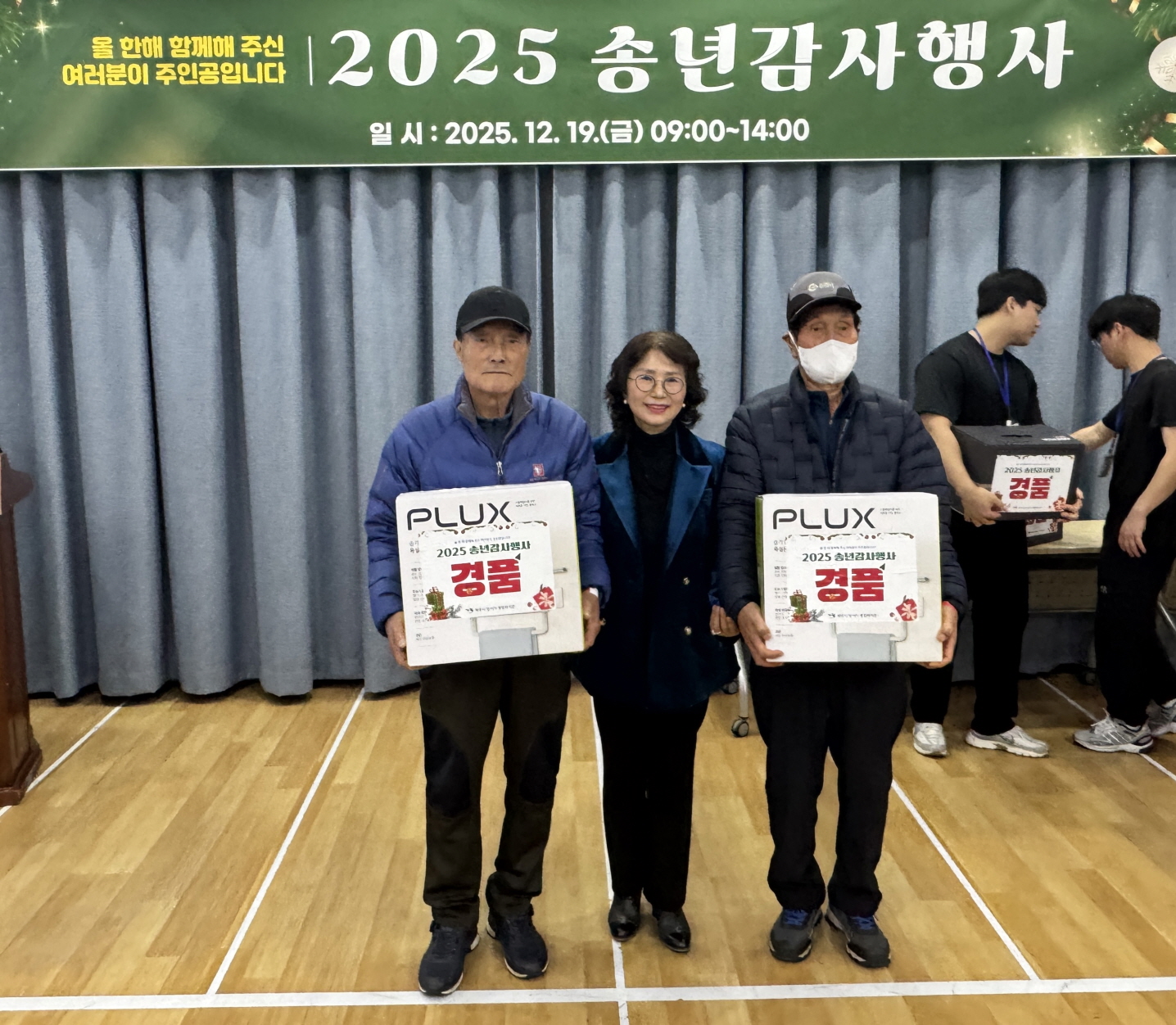 2025 송년감사행사에서 경품 당첨자 두 명이 대형 경품 상자를 들고 나란히 서 있고, 관계자가 옆에서 함께 포즈를 취하며 당첨을 기념해 촬영하고 있다.