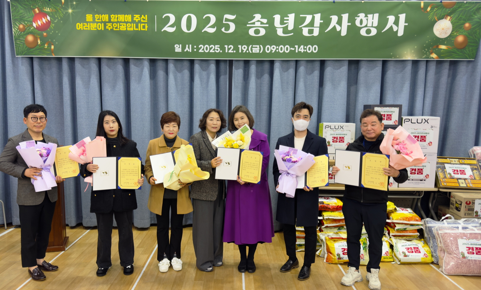 2025 송년감사행사에서 유공자 표창 수상자들이 상장과 꽃다발을 들고 기념촬영을 하며, 옆에 경품과 후원물품이 진열되어 있다.