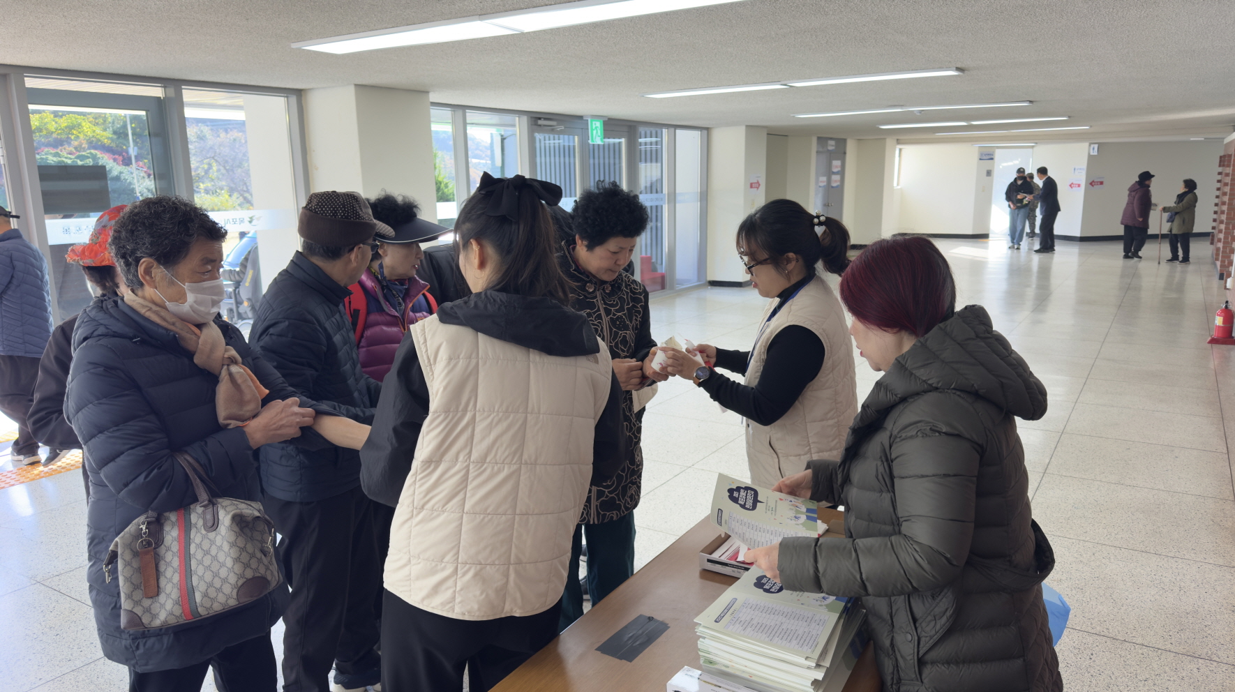 지역사회 공헌활동의 일환으로 실내 로비 안내 테이블에서 직원들이 참가자에게 안내물과 입장권을 나눠주며 친절히 안내하는 모습. 참가자들이 줄을 서서 순서대로 접수하고 있으며, 책상 위에는 행사 안내책자와 자료가 놓여 있다.