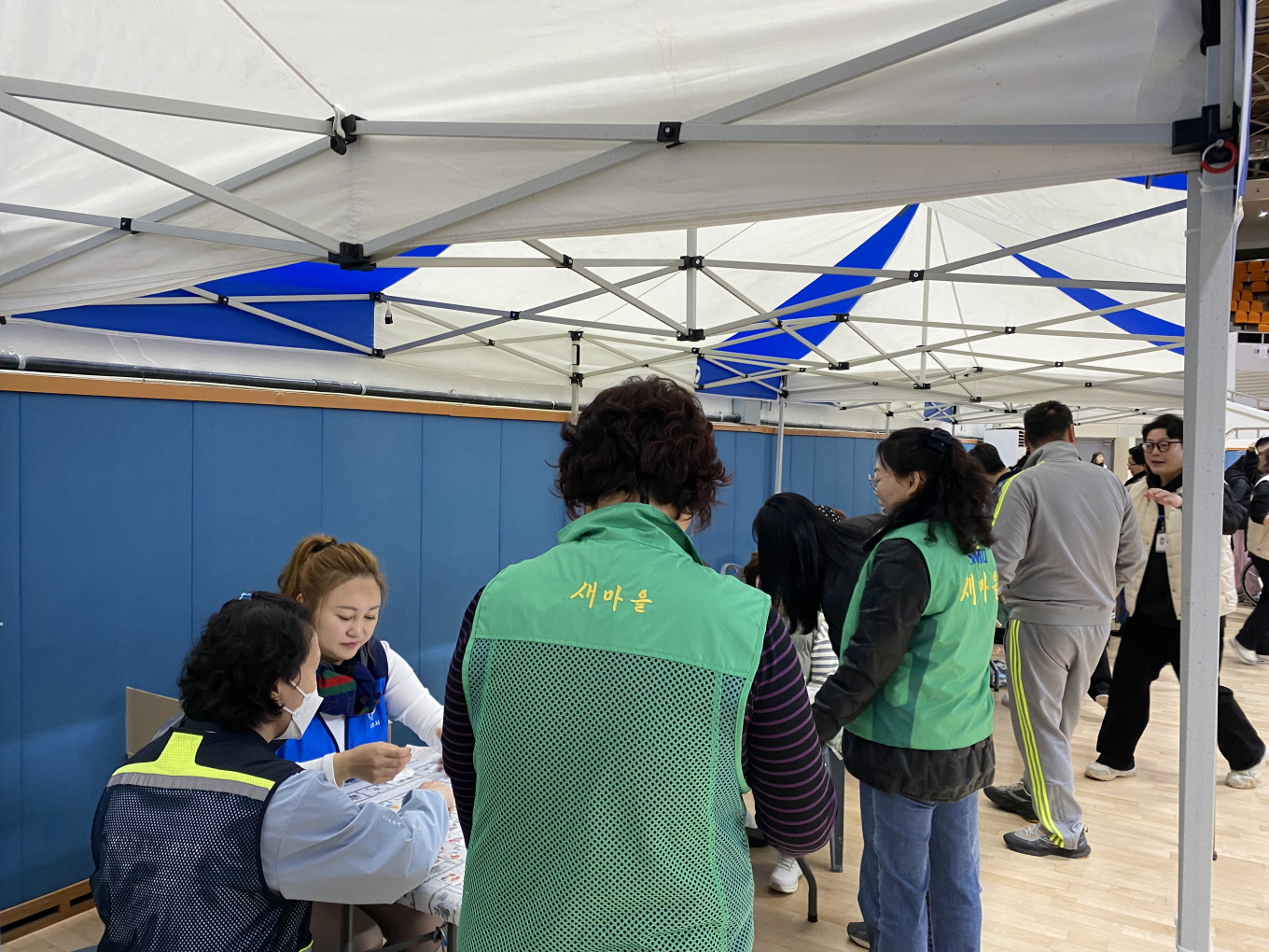 지역사회 공헌활동으로 행사장 천막 부스에서 녹색 조끼를 입은 하당동 새마을부녀회 봉사자들이 참가자 접수와 안내를 돕는 모습. 파란색 조끼를 입은 ‘목포를 만드는 사람들’ 봉사자도 함께 참여해 부스 내에서 협력하고 있으며, 주변으로는 참가자들이 오가고 있다.