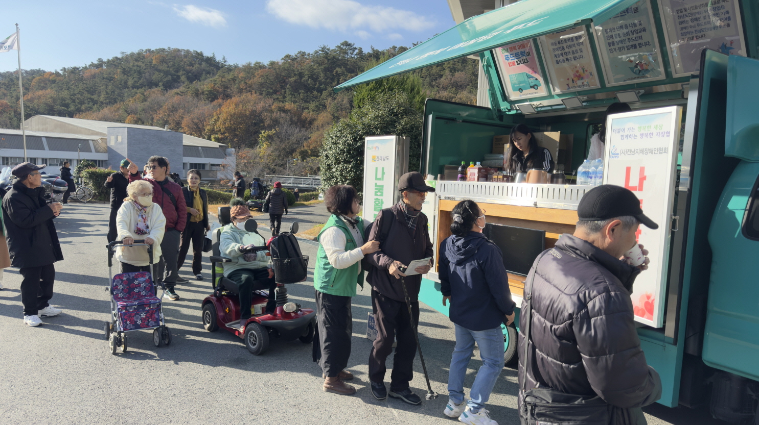 지역사회 공헌활동으로 운영된 야외 푸드트럭 앞에서 참가자들이 줄을 서서 음료와 간식을 받기 위해 기다리는 모습. 보행보조기나 전동 이동기기를 이용하는 장애인 참가자들도 함께 줄을 서 있으며, 푸드트럭 안에는 봉사자가 주문을 받고 있다.
