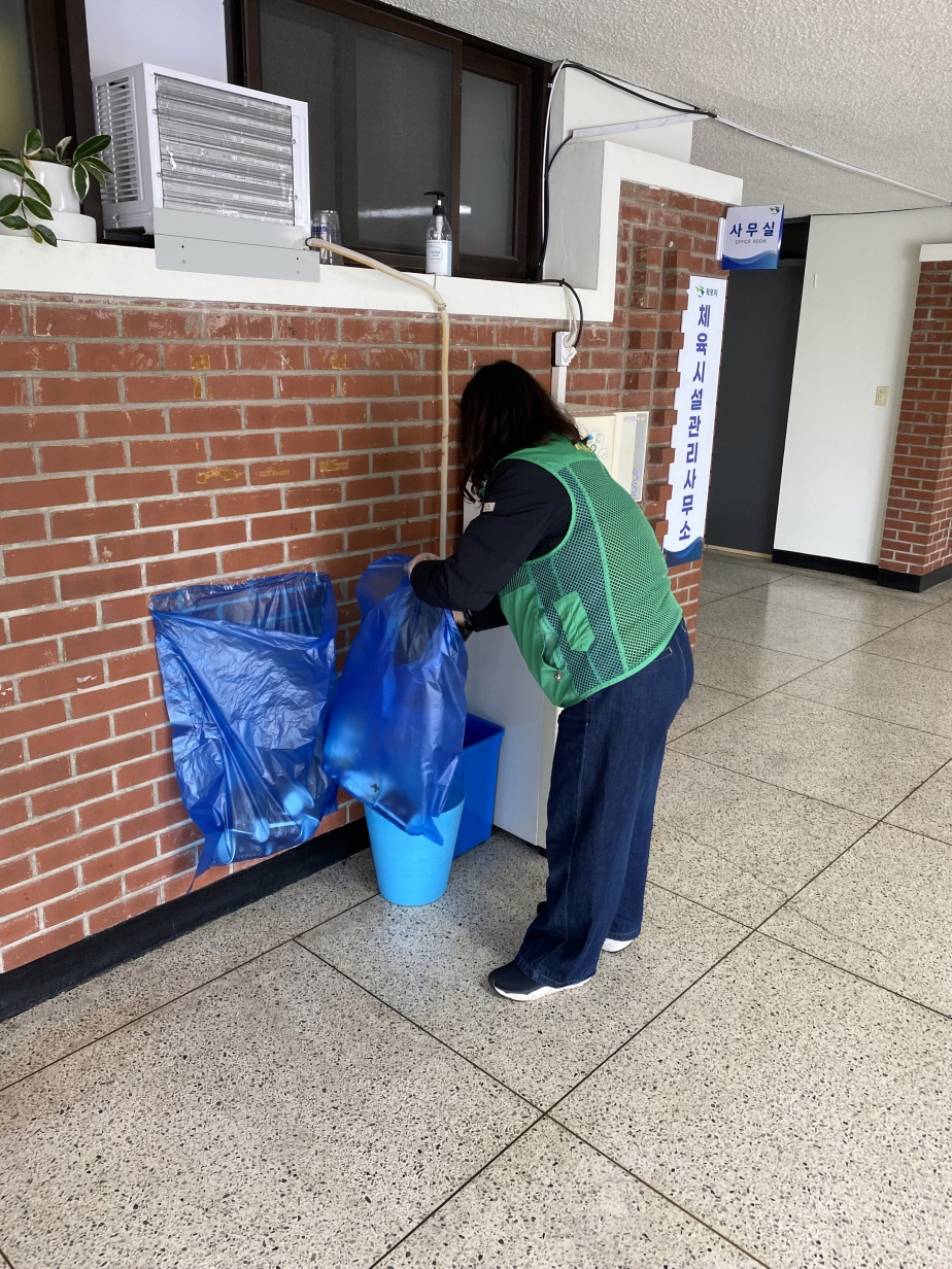 지역사회 공헌활동의 일환으로 실내 복도에서 녹색 조끼르 입은 하당동 새마을부녀회 봉사자가 쓰레기통의 파란 비닐봉투를 교체하며 주변 환경을 정리하는 모습. 행사 종료 후 복도 환경을 깨끗이 유지하기 위해 정리정돈을 수행하고 있다.