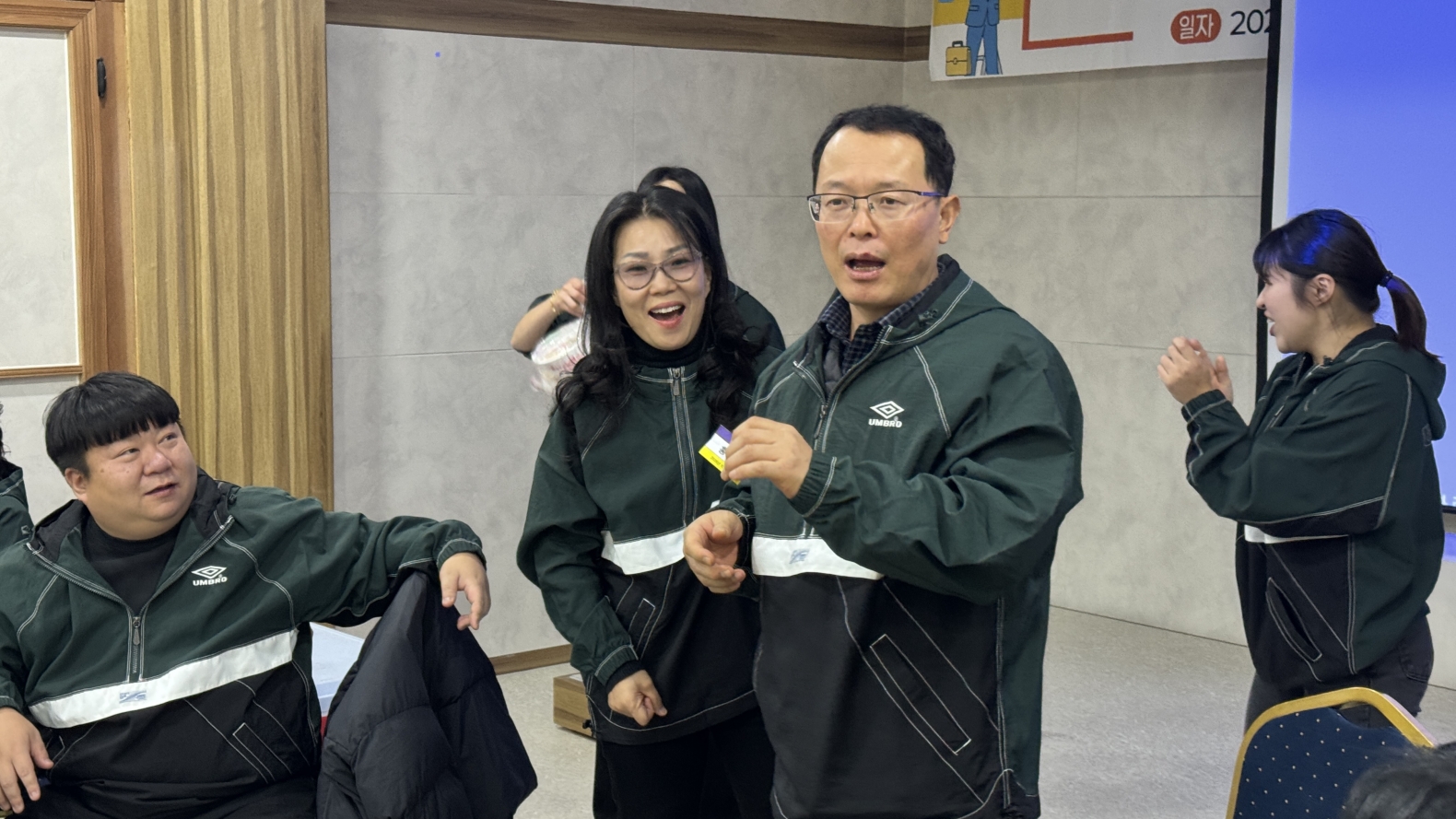 전 직원 소통의 시간. 한 남성 직원이 마이크 없이 동료들 앞에서 밝은 표정으로 소감을 발표하고 있으며, 주변 동료들이 박수를 치며 경청하고 있다.