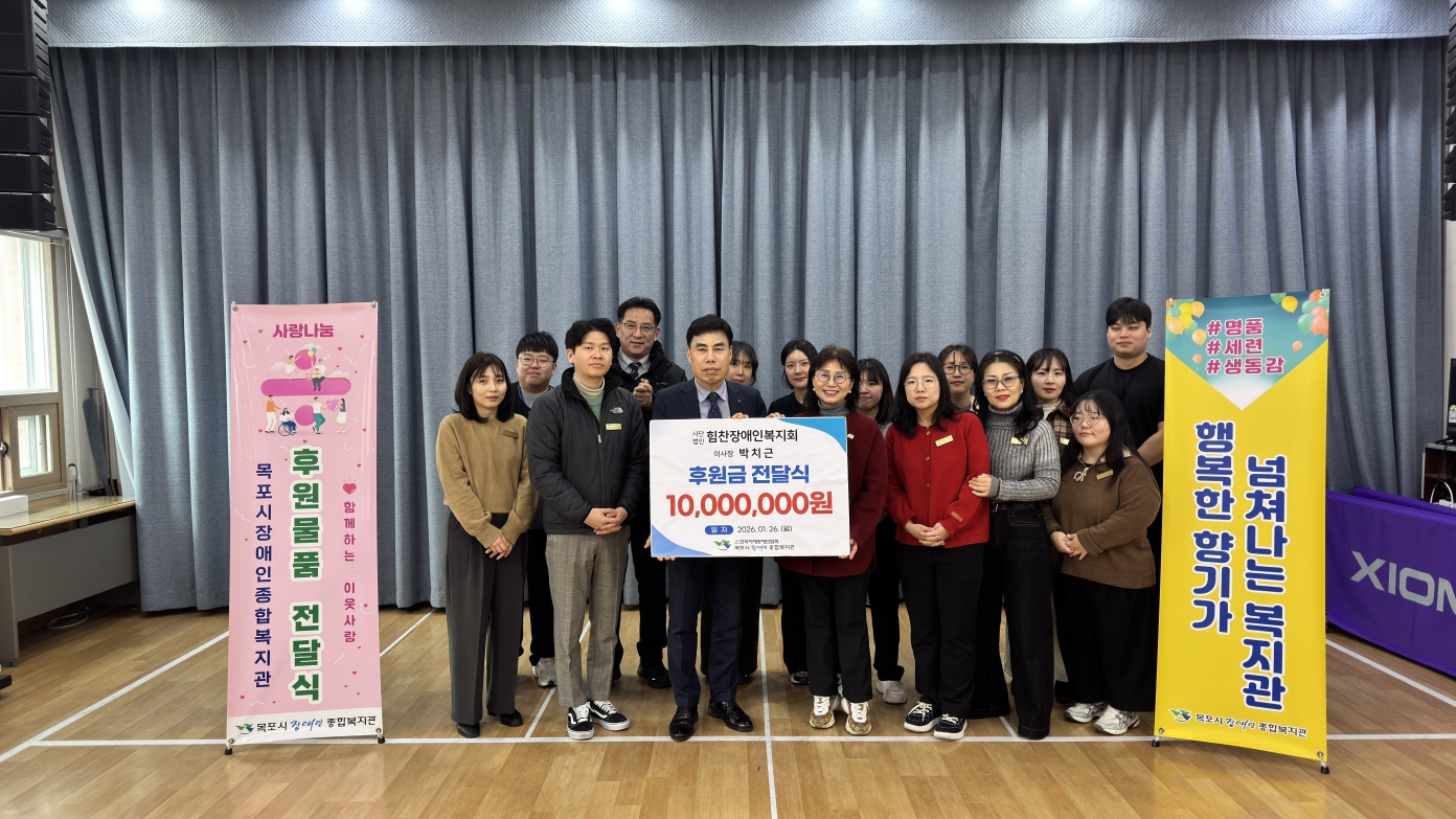 사단법인 힘찬장애인복지회 박치근 이사장의 후원금 전달식 단체 기념사진. 무대 중앙에서 박치근 이사장과 목포시장애인종합복지관 관계자들이 '후원금 10,000,000원'이라고 적힌 대형 판넬을 함께 들고 기념 촬영을 하고 있습니다. 양옆에는 '후원 물품 전달식'과 '행복한 향기가 넘쳐나는 복지관'이라는 문구가 적힌 배너가 세워져 있습니다.