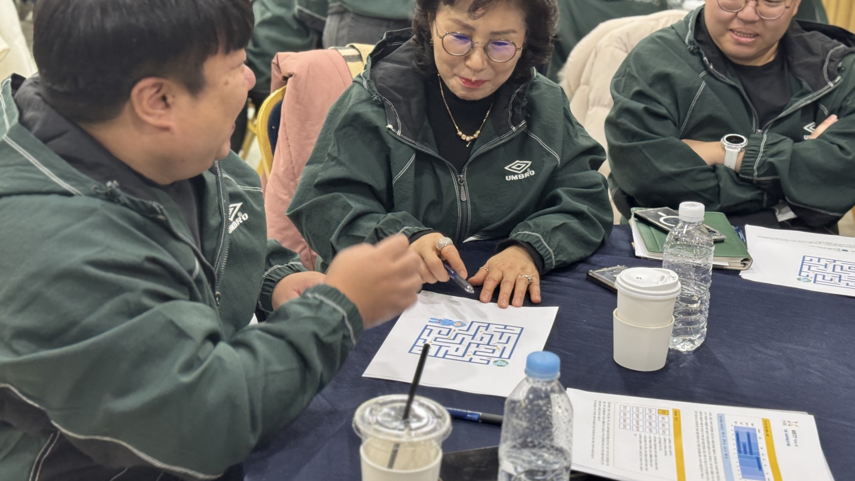직원들이 조직 활성화를 위한 미로 찾기 활동지를 함께 해결하며 밝게 웃고 있는 모습. 두 명이 머리를 맞대고 협력하며 소통하고 있다.
