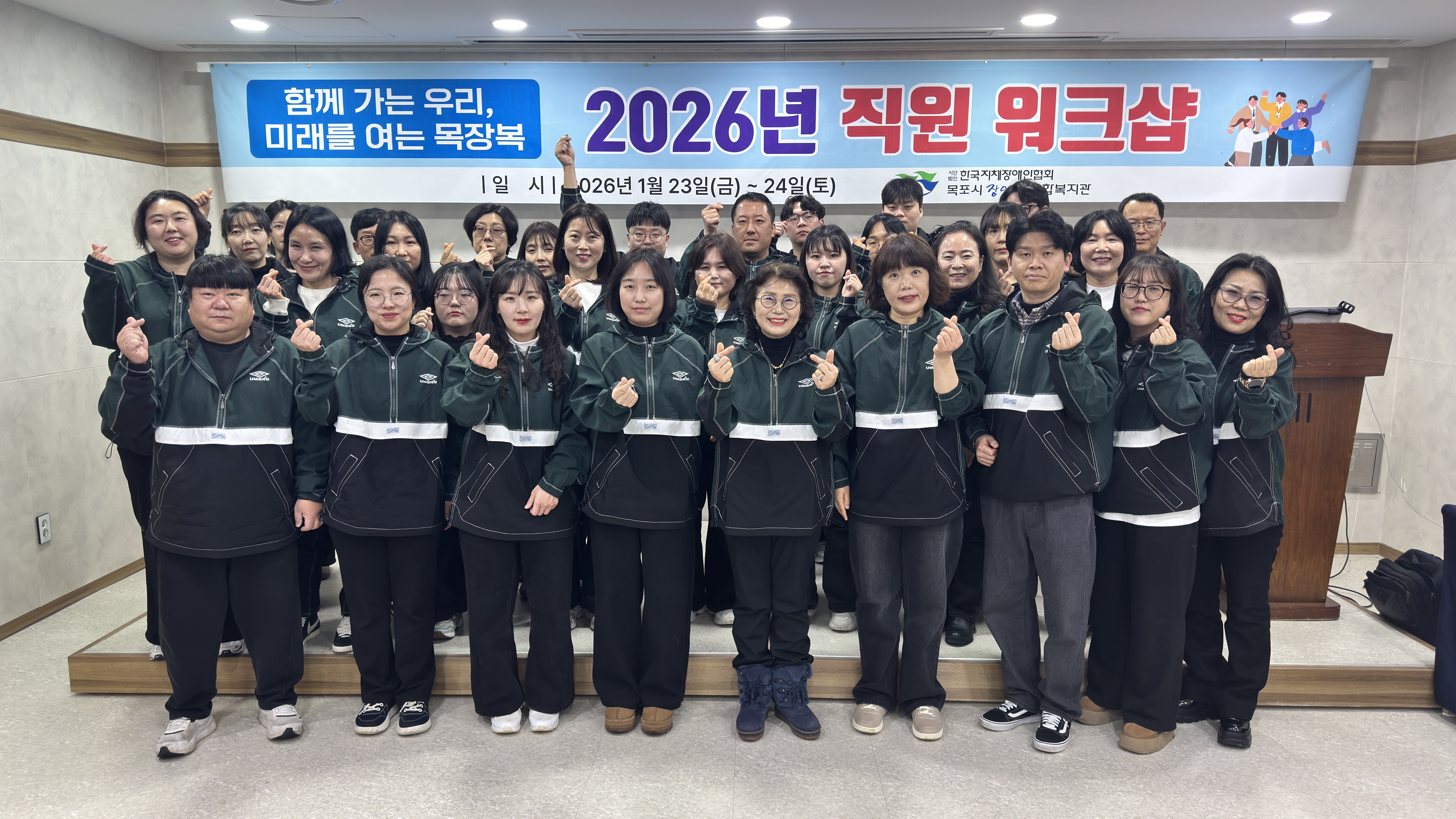 2026년 전 직원 역량강화 워크숍 단체 사진. '함께 가는 우리, 미래를 여는 목장복!' 현수막 아래에서 초록색 단체복을 입은 직원들이 손하트 포즈를 취하며 밝게 웃고 있다.