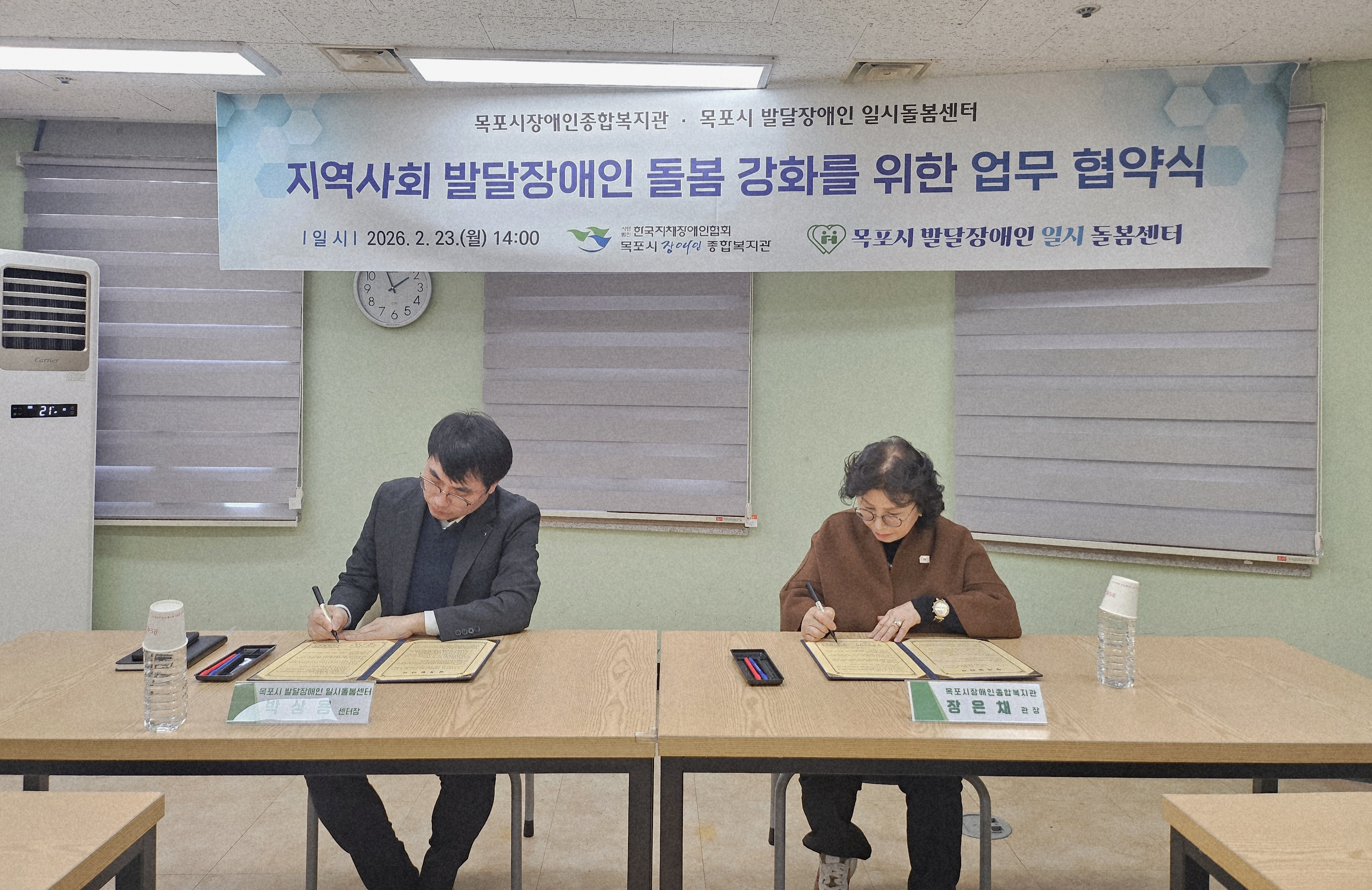 목포시 발달장애인 일시돌봄센터 박성용 센터장과 목포시장애인종합복지관 장은채 관장이 책상에 나란히 앉아 업무 협약서에 서명을 하고 있는 모습입니다. 두 사람 앞에는 각각 이름표가 놓여 있으며, 진지한 표정으로 서류에 집중하고 있습니다. 배경에는 '지역사회 발달장애인 돌봄 강화를 위한 업무 협약식'이라는 문구와 함께 일시(2026. 2. 23. 월 14:00) 및 양 기관의 로고가 포함된 현수막이 걸려 있습니다.