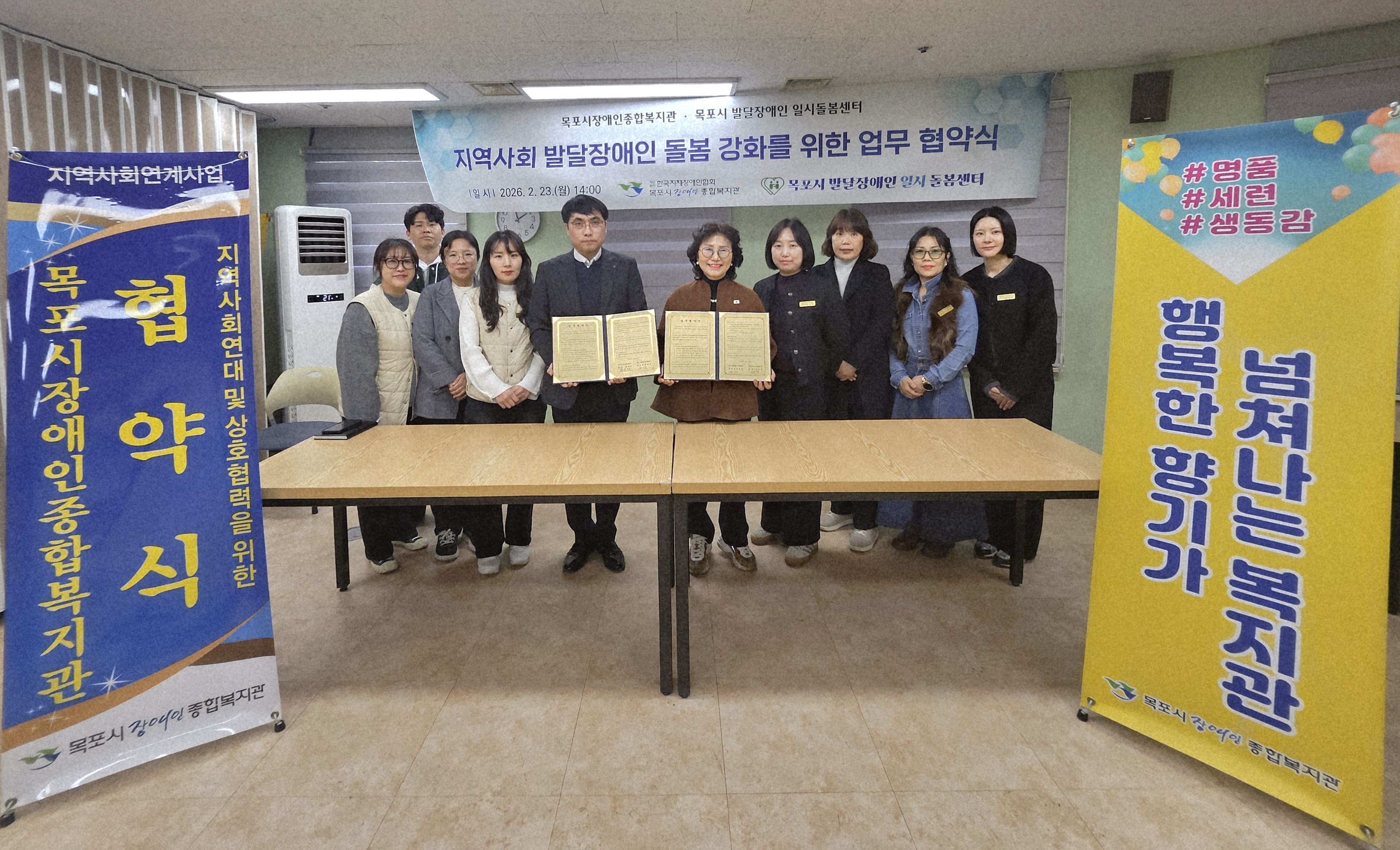 2026년 2월 23일 오후 2시, 목포시장애인종합복지관과 목포시 발달장애인 일시돌봄센터가 체결한 '지역사회 발달장애인 돌봄 강화를 위한 업무 협약식' 기념 촬영 사진입니다. 중앙에는 양 기관의 대표인 박성용 센터장과 장은채 관장이 서명된 협약서를 나란히 들고 있으며, 그 좌우로 8명의 관계자들이 함께 서서 정면을 응시하고 있습니다. 배경에는 협약식 명칭이 적힌 현수막이 걸려 있고, 좌측에는 '협약식' 문구가 적힌 파란색 배너가, 우측에는 '넘쳐나는 복지관 행복한 향기'가 적힌 노란색 배너가 세워져 있습니다.