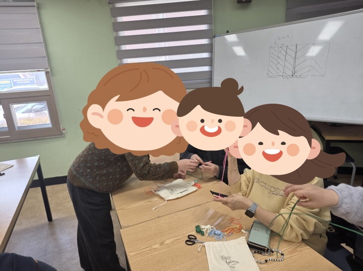 강사와 교육생들이 강의실 책상에 둘러앉아 실이나 끈을 활용한 수공예(매듭 만들기 등) 실습을 진행하고 있는 모습. 강사가 교육생의 작업을 가까이서 도와주고 있으며, 책상 위에는 가위, 알록달록한 실 꾸러미, 작은 에코백 등의 실습 도구들이 놓여 있다. 배경의 화이트보드에는 매듭법이나 도안으로 보이는 그림이 그려져 있다.