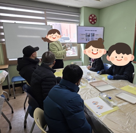 교육장에서 강사가 수강생들에게 교육을 진행하는 모습. 연두색 상의를 입은 강사가 화이트보드와 모니터 앞에 서서 교구(투명 용기)를 들어 보이며 설명하고 있고, 테이블에 둘러앉은 4명의 수강생이 강사를 집중해서 보고 있다. 테이블 위에는 교육용 키트와 안내문 등이 놓여 있으며, 오른쪽 벽면 모니터에는 교육 자료 화면이 띄워져 있다.