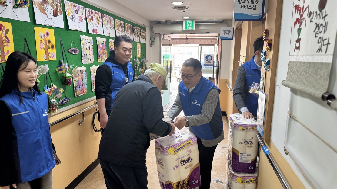 목포시장애인종합복지관 복도에서 목포토박이봉사단 회장이 노란 모자를 쓴 이용고객에게 후원 물품인 대형 화장지를 직접 전달하고 있는 모습. 회장은 파란색 단체 조끼를 입고 있으며, 주변에는 다른 봉사자들이 물품 전달을 돕고 있음. 복도 왼쪽 초록색 게시판에는 장애인들이 그린 알록달록한 그림들이 전시되어 있고, 우측 상단에는 '방과 후 활동 서비스실' 표지판이 보임.