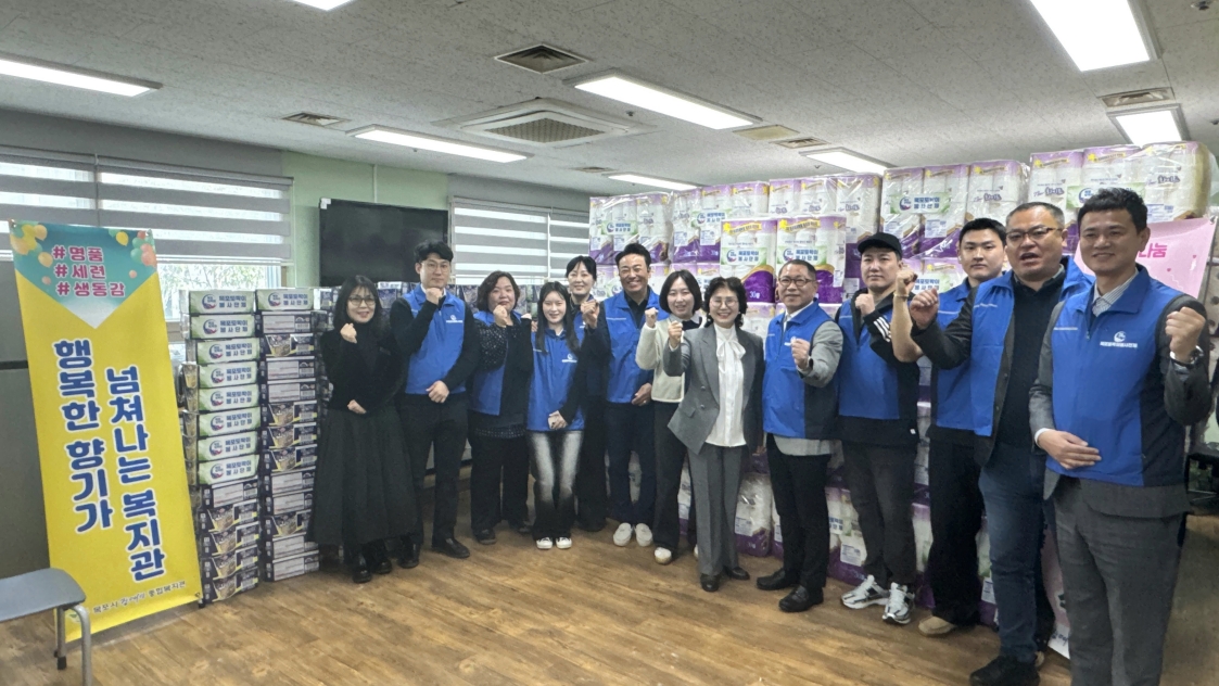 목포토박이봉사단 회원들과 복지관 관계자 약 15명이 후원 물품 앞에서 단체로 파이팅 포즈를 취하며 기념 촬영을 하는 모습. 왼쪽에는 '명품, 세련, 생동감, 넘쳐나는 복지관, 행복한 향기가'라고 적힌 노란색 배너가 있고, 뒤쪽으로는 기탁된 대량의 화장지와 상자들이 가득 쌓여 있음.