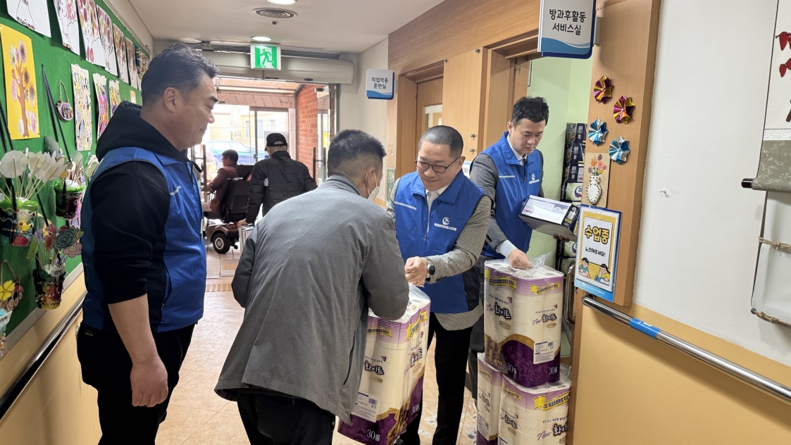 목포시장애인종합복지관 복도에서 목포토박이봉사단 회장이 복지관 이용고객에게 후원 물품을 전달하는 모습. 회장은 파란색 조끼를 입고 있으며, 옆에 선 다른 봉사자는 상자에 담긴 세제류 물품을 준비하고 있음. 복도 벽면에는 '방과 후 활동 서비스실', '직업적응 훈련실' 표지판이 보이고, 그 옆으로 대량의 화장지 꾸러미가 쌓여 있음.