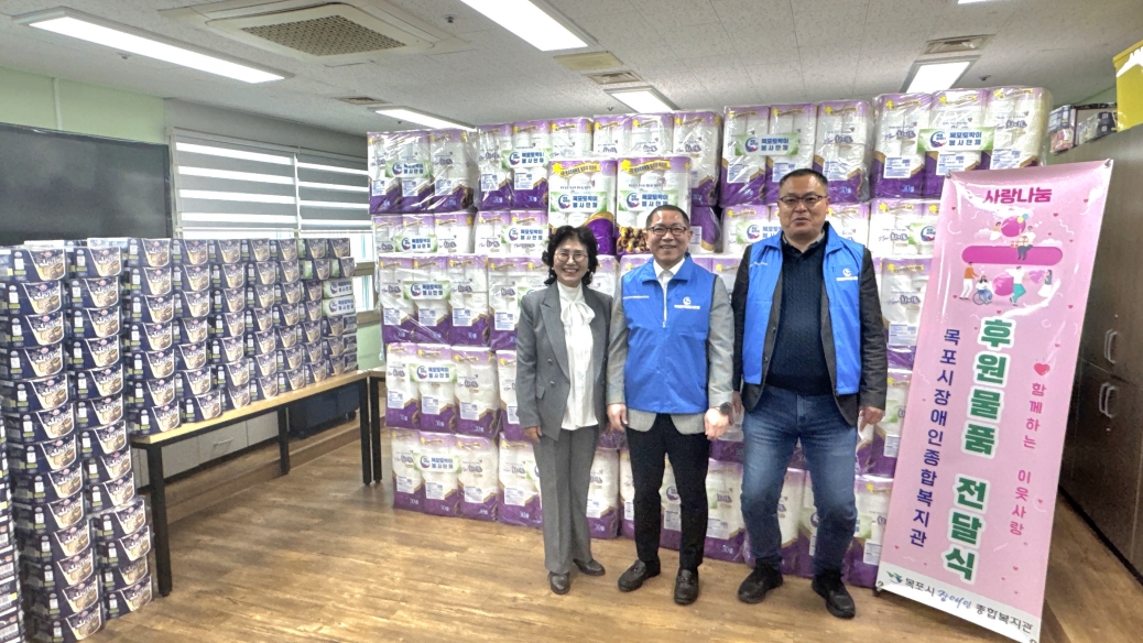 목포시 장애인 종합복지관 내부에서 목포토박이봉사단 관계자 3명이 300만 원 상당의 후원 물품(화장지, 세제 등) 앞에서 기념 촬영을 하고 있는 모습. 오른쪽에는 '사랑 나눔 후원 물품 전달식, 함께하는 이웃사랑, 목포시장애인종합복지관'이라고 적힌 핑크색 배너가 세워져 있고, 배경에는 기부된 화장지가 높게 쌓여 있음.