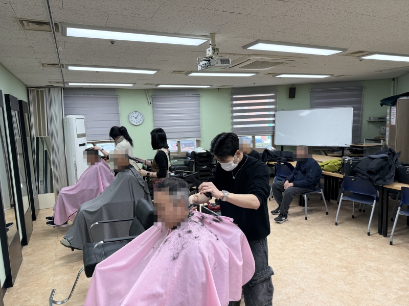 실내 교육실 또는 실습실에서 세 명의 이용자가 의자에 앉아 이발 서비스를 받고 있고, 마스크를 착용한 봉사자들이 이발기(클리퍼)와 빗을 사용하여 정성스럽게 머리카락을 다듬고 있는 모습.