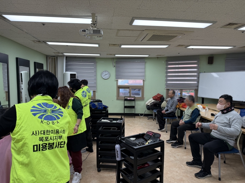 (사)대한미용사회 목포시지부 미용봉사단 활동 모습. 전경의 봉사자가 입은 형광색 조끼 뒷면에 'K·O·B·A 사) 대한미용사회 목포시지부 미용봉사단' 문구와 로고가 명확히 보인다. 배경으로는 미용 트레이들과 머리 손질을 기다리며 앉아 있는 이용고객들이 보인다.