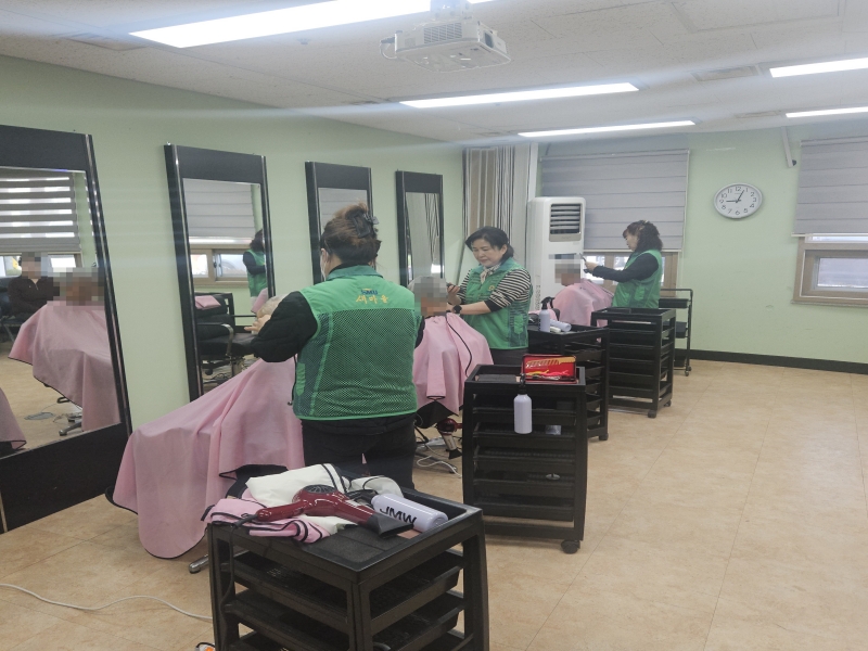 녹색 조끼를 입은 봉사자들이 실내에서 노인들에게 이용 및 미용 봉사를 하고 있는 모습